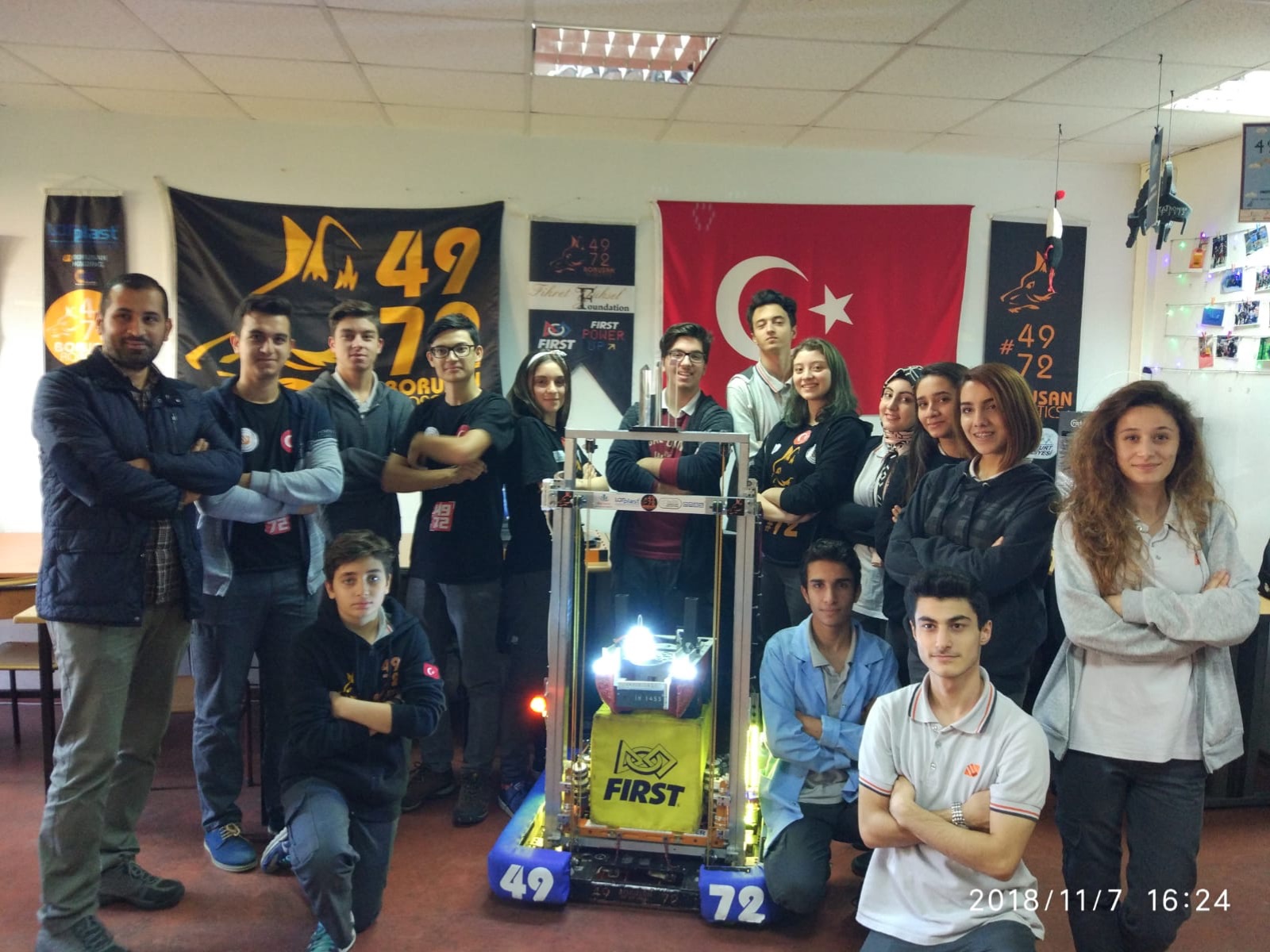 Takım, okul atölyesinde robotun etrafında; arkada Türk bayrağı ve takım flamaları